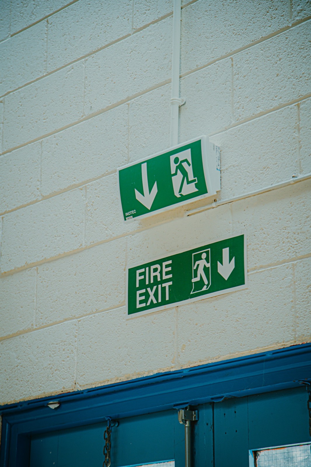 Green fire exit signs mounted on the painted brickwork of a British building above a blue door.