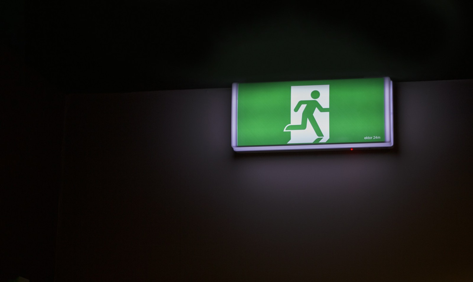 An illuminated green fire exit sign showing the running man symbol, lit in a dark room.